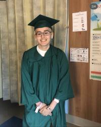 Image of Carlos Romero in green graduation cap and gown, standing in his hospital room.
