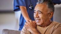 Man sitting on a couch smiling while a healthcare provider stands behind him with hands on his shoulders.