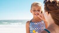 Photo of a woman applying sunscreen on a little girl in a bathing suit with the beach in the background.