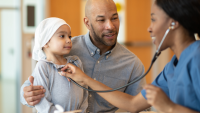 A doctor checking a small child with a stethoscope while the child's parent stands beside them and holds them.